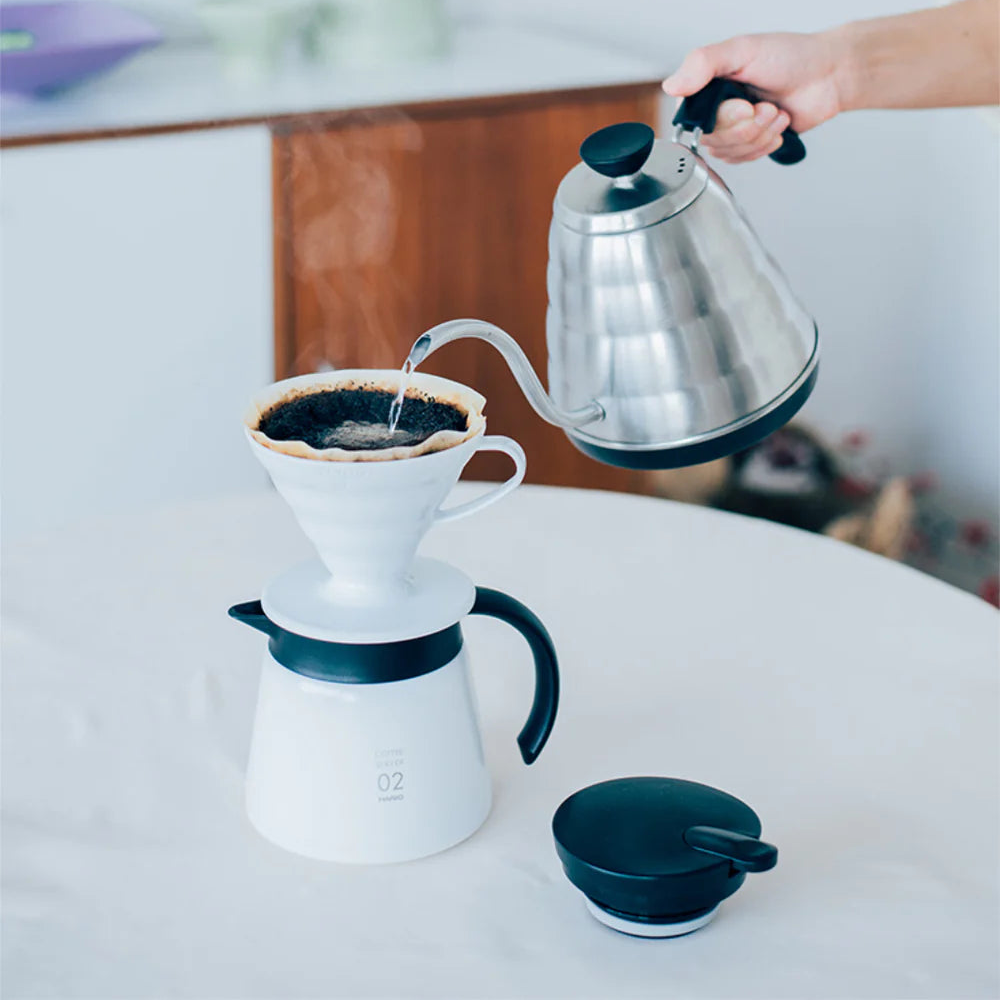 Person pouring coffee from a silver kettle into a white coffee maker on a light surface.